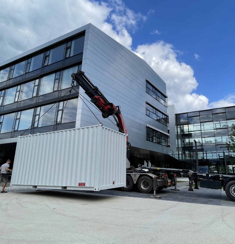 Kran hebt einen weißen Container vor dem Gebäude eines Rechenzentrums. Im Hintergrund sieht man den Himmel, der blau mit Wolken ist.