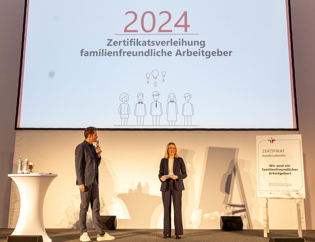 Moderator und Bernadett Humer, Sektionschefin fuer Famiie und Jugend, stehen auf Buehne und teilen Zertifikate aus